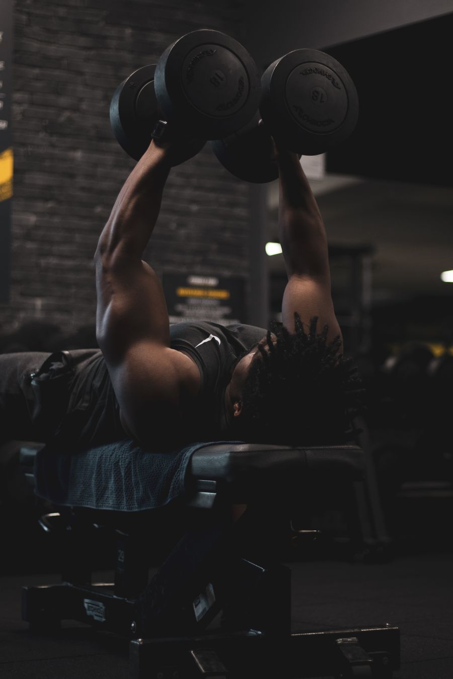 A man on a bench in a dim gym, pressing two dumbbells overhead.